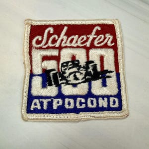 May include: A square embroidered patch featuring a red top section with the white cursive text Schaefer, a central white 500 with a black race car graphic, and a blue bottom section with the white text AT POCONO, all bordered in white thread.