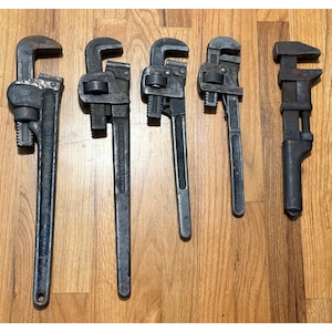 May include: A collection of five dark grey metal pipe wrenches of varying sizes, arranged in a row. Each tool features a serrated jaw, an adjustable nut mechanism, and a long handle, showing signs of wear and metallic texture throughout.