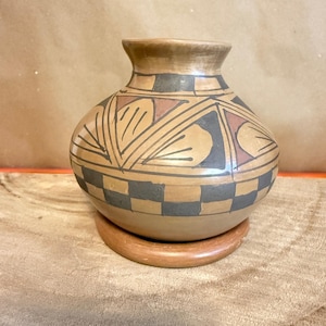 May include: A handcrafted, round pottery vase with a wide body and a narrow neck. The vase features geometric designs in black, brown, and red against a tan background. It sits on a circular, wooden base, showcasing traditional craftsmanship.