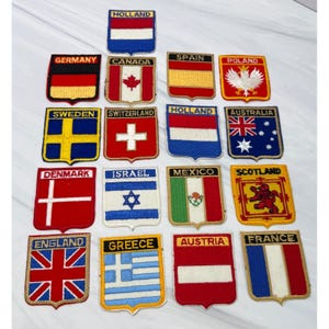 Puede incluir: Una colección de diecisiete parches bordados rectangulares, cada uno con una bandera nacional y el nombre del país correspondiente en texto negro. Los parches están dispuestos en una cuadrícula y muestran varios diseños coloridos, incluidos símbolos como la hoja de arce canadiense, el águila polaca y el león escocés.
