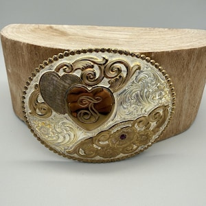 May include: Oval-shaped silver and gold belt buckle with an ornate design. Features two heart shapes, one silver and one gold, with a decorative letter. The buckle has a detailed floral and scroll pattern, with a small red jewel accent.