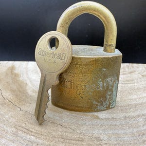 May include: A vintage brass padlock with a matching key. The key is inserted into the lock, which has the word "American" and "U.S.A." engraved on it. The lock is a warm, aged brass colour.