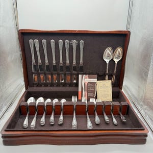 May include: A wooden silverware chest with a dark brown finish, open to reveal a complete set of silver-colored flatware. The set includes knives, forks, spoons, and serving spoons, all neatly arranged in a dark brown felt-lined interior. The silverware has a decorative floral pattern.