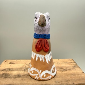 May include: A handcrafted ceramic bird figurine with a light purple head, brown beak, and blue collar. The body is tan with a red bow tie and white painted details. The figurine is on a wooden surface.