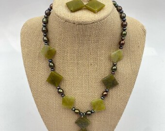 May include: A necklace and earring set featuring olive green square-cut stones and dark pearl beads. The necklace has a diamond-shaped pendant. The earrings have matching square stones. The jewelry is displayed on a tan mannequin.