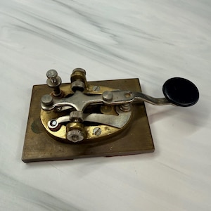 May include: A vintage telegraph key mounted on a rectangular brass base. The device features a silver-toned metal lever with a circular black knob at one end, brass adjustment screws, and a central spring mechanism for manual signal transmission.