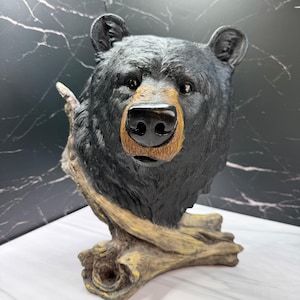 May include: A black bear head sculpture featuring textured fur, a prominent snout with a tan-coloured muzzle, and a glossy black nose. The bear is depicted emerging from a textured, light brown wooden log base with visible knots and grain details.
