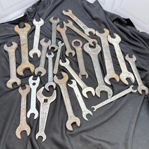 Op de afbeelding: Een verzameling diverse metalen moersleutels van verschillende groottes en vormen, met steek- en ringsleutelontwerpen. De gereedschappen vertonen tekenen van slijtage, waaronder roest en oppervlakte-oxidatie, verspreid over een donkere stof.