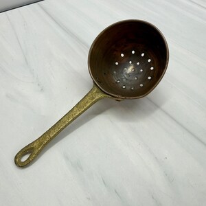May include: A copper skimmer with a round, perforated bowl and a long, textured brass handle. The bowl features a series of small, evenly spaced holes for draining liquids, while the handle has a loop at the end for hanging storage.