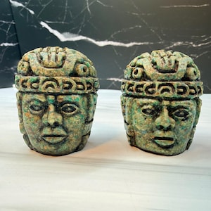 May include: Two carved stone sculptures depicting human heads with detailed headdresses. The figures feature a mottled green and brown texture, prominent facial features, and a structured, layered crown design across the top of each head.