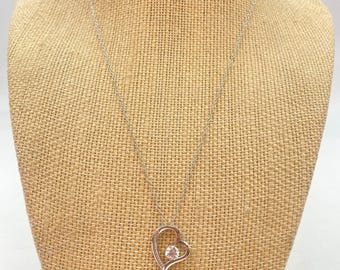 May include: A silver-toned necklace with a heart-shaped pendant. The pendant features a clear, round gemstone set within the heart's outline. The necklace is displayed on a tan fabric background.