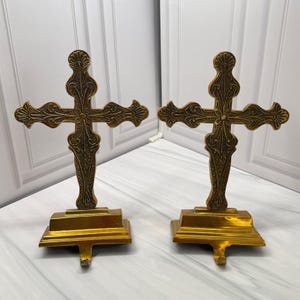 Könnte beinhalten: Ein Paar goldfarbene Metall-Strumpfhalter, jeweils mit einem verzierten Kreuzdesign und detaillierten Prägungen. Die Kreuze sind auf rechteckigen, gestuften Sockeln montiert, wobei ein gebogener Haken an der Vorderseite jedes Sockels hervorsteht.