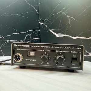May include: A grey Kenwood Phone Patch Controller PC-1A featuring a circular microphone input, a small analog meter labeled Moni/Null, two rotary knobs for RX Gain and TX Gain marked from 0 to 10, and a black toggle switch for Patch On/Off.