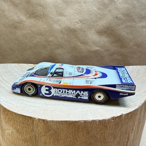 May include: A die-cast model race car featuring a white and blue livery with the number 3 on the side. The car displays Rothmans Porsche branding, Shell logos, and Dunlop text. It has gold-colored wheels and a low-profile aerodynamic body design.