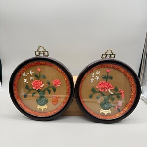 May include: Two round framed wall art pieces. Each features a floral arrangement in a vase, with red and pink flowers. The frames are dark wood with gold accents. Chinese characters are visible on each piece.