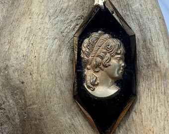 May include: A diamond-shaped pendant featuring a metallic gold-toned profile portrait of a person with curly hair set against a smooth black background. The pendant has a dark metallic frame and a silver-toned metal bail at the top for hanging.