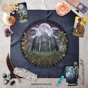 May include: A dark blue fabric tapestry with a circular design featuring three standing stones, a full moon, and a crescent moon. The design is surrounded by a border of repeating crescent moons. The tapestry has the text "brightarrow.etsy.com/" at the bottom.