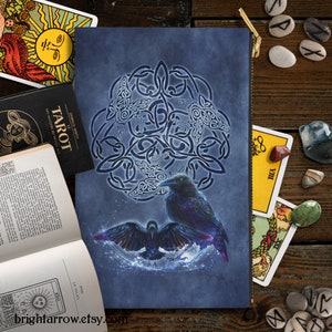 May include: A blue zippered pouch featuring a Celtic knot design and two black ravens. The ravens are surrounded by a glowing circle of light. The pouch has a zipper at the top and a gold zipper pull.