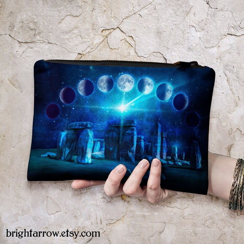 May include: A blue and black zippered pouch with a cosmic design featuring a crescent moon, stars, and a stonehenge-like structure. The pouch is made of canvas and has a zipper closure.