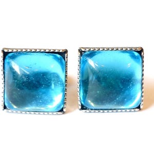 Könnte beinhalten: Zwei quadratische, hellblaue Ohrringe mit silberfarbenem, strukturiertem Rand. Die Ohrringe haben ein durchscheinendes, glasartiges Aussehen und sind vor einem weißen Hintergrund platziert. Es handelt sich um ein Schmuckstück.