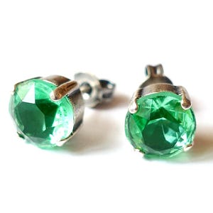 Può includere: Un paio di orecchini a bottone con pietre preziose rotonde, verde smeraldo, incastonate in metallo argentato. Le pietre sfaccettate brillano su uno sfondo bianco, evidenziando il colore verde.
