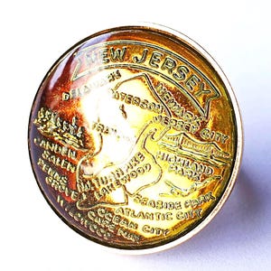 Könnte beinhalten: Ein runder, goldfarbener Ansteckpin mit einer Karte von New Jersey, einschließlich Städten wie Camden und Atlantic City. Der Pin hat eine klare, gewölbte Oberseite und einen goldenen Rand. Der Text "NEW JERSEY" ist oben gewölbt.