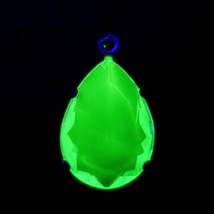 Könnte beinhalten: Ein leuchtend grüner, tropfenförmiger Kristallanhänger mit facettiertem Design. Er hat eine kleine blaue Schlaufe zum Aufhängen und dunkelblaue Akzente. Der Hintergrund ist schwarz.