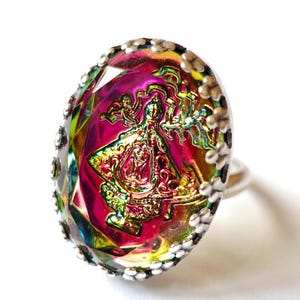 Könnte beinhalten: Ovaler Ring mit silbernem Band. Der Ring hat einen großen, facettierten, irisierenden Stein mit einem zentralen Design in Rosa, Gold und Grün. Der Stein ist von einer silbernen Lünette mit kleinen, dekorativen Akzenten umgeben.