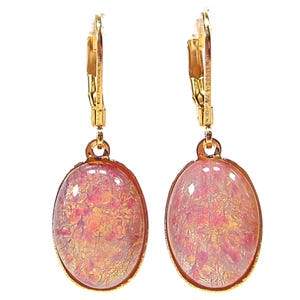 May include: A pair of gold-coloured leverback earrings. Each earring features an oval-shaped, pink and gold-coloured stone. The stones have a textured, iridescent appearance, with a mix of pink and gold hues. The earrings are set against a white background.