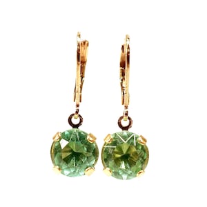 May include: A pair of gold-colored leverback earrings, each featuring a round, faceted, light green gemstone. The gemstones are set in a gold-colored bezel. The earrings are displayed against a white background.