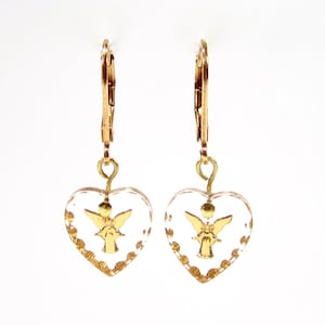 Puede incluir: Pendientes de corazón dorados con un diseño de cristal transparente y un ángel dorado dentro de cada corazón.