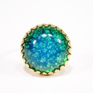 May include: A gold-coloured ring with a striking blue-green centre. The centre has a textured, speckled appearance, and the gold band features a delicate, repeating pattern around the edge. The ring is set against a white background.