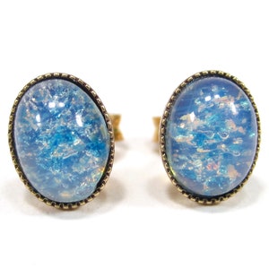 Op de afbeelding: Een paar ovale oorbellen met een blauwe, iriserende glazen cabochon, gezet in een gouden metalen bezel.