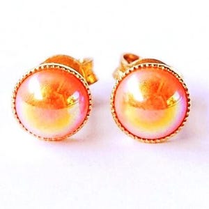 Può includere: Un paio di orecchini a bottone color oro. Ogni orecchino presenta una pietra rotonda, iridescente, arancione-rosa, incastonata in una lunetta dorata dettagliata. Gli orecchini sono presentati su sfondo bianco.