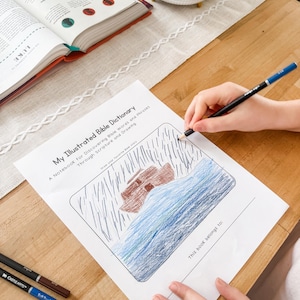 Pode incluir: Um desenho infantil de um barco sobre um mar azul, colorido com lápis, numa página de "My Illustrated Bible Dictionary". A página está numa mesa de madeira ao lado de um livro aberto e de um recipiente branco com lápis de cor.
