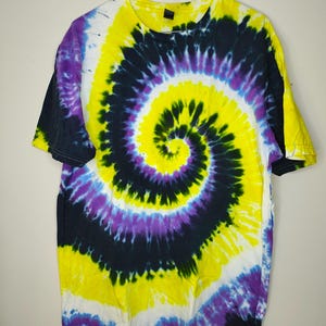 Puede incluir: Una camiseta de manga corta con cuello redondo que presenta un vibrante patrón de espiral tie-dye. El diseño incorpora remolinos audaces de amarillo brillante, negro intenso, morado real y blanco, creando un gráfico dinámico en la prenda.