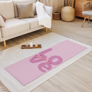 Puede incluir: Una esterilla de yoga rosa con las palabras "LOVE you" en una fuente rosa más oscura. La esterilla es rectangular y está sobre una alfombra blanca. El fondo incluye un sofá beige, un armario de madera y una cesta tejida.
