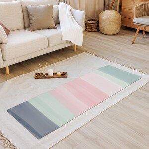 Puede incluir: Una esterilla de yoga rectangular con un diseño de acuarela. La esterilla presenta rayas horizontales grises, verde menta, rosas y azul claro. La esterilla está sobre una alfombra blanca en una habitación con un sofá y muebles de madera.