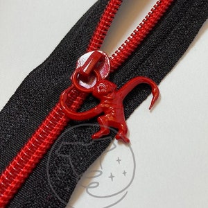 Op de afbeelding: Een zwarte rits met rode tanden en een rode apenritstrekker. De rits is gedeeltelijk open, waardoor de rode tanden op de zwarte stof zichtbaar zijn. De apenritstrekker is felrood.