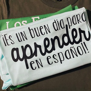 May include: A stack of light blue and green t-shirts with the text "¡Es un buen día para aprender en español!" printed in black.