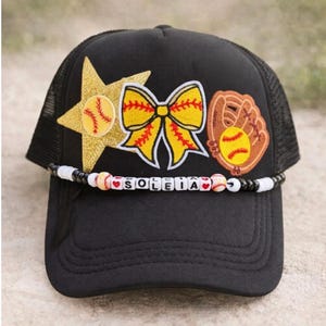 May include: Black baseball cap with a mesh back, adorned with softball-themed patches. Features a gold star, yellow bow, and a softball glove. A beaded band spells out "SOLEIA".