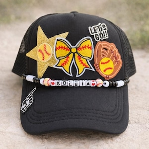 May include: Black baseball cap with a mesh back, decorated with a gold star, yellow bow, and baseball glove patches. The cap features the text "LET'S GO!!" and a beaded band with the name "SOLEIA".