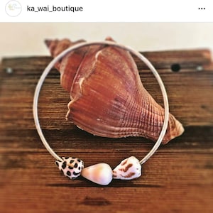 Może przedstawiać: Srebrny naszyjnik typu choker z trzema muszlami. Muszle są białe, brązowe i beżowe, z unikalnymi wzorami. Naszyjnik jest prezentowany na drewnianej powierzchni, z dużą muszlą w tle.