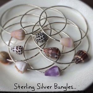 May include: A collection of sterling silver bangles, each adorned with a unique seashell. The shells vary in color, including white, brown, and purple, and are strung on the silver bands. The text "Sterling Silver Bangles..." is visible.