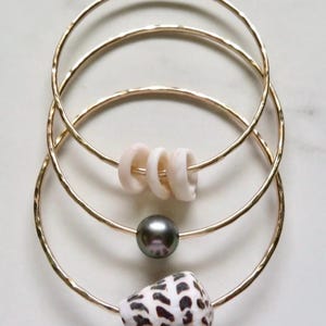 Puede incluir: Cuatro brazaletes dorados, cada uno con un encanto único. Un brazalete presenta una perla negra, otro tiene una concha marina y un tercero tiene cuentas de concha blanca. Los brazaletes están dispuestos en una pila.