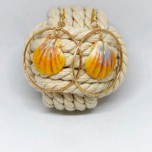 Peut inclure: Boucles d'oreilles créoles dorées avec des pendentifs en forme de coquillage. Les coquillages sont peints en jaune, orange et blanc. Les boucles d'oreilles sont présentées sur un fond de corde beige. Diamètre des créoles : environ 5 cm.