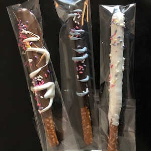 May include: Three chocolate-covered pretzel rods in clear cellophane wrappers. One is dipped in dark chocolate with white drizzle and sprinkles. Another is dipped in dark chocolate with blue drizzle and sprinkles. The third is dipped in white chocolate with sprinkles.