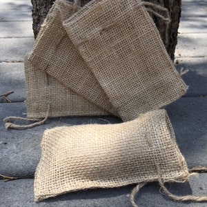 Op de afbeelding: Verschillende kleine, rechthoekige jute zakken met trekkoord sluitingen. De zakken hebben een natuurlijke bruine kleur en zijn in een groep gerangschikt, met &eacute;&eacute;n zak plat en de andere tegen een boomstam. De zakken zijn gemaakt van een grof geweven stof.