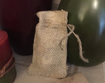 May include: A small, beige burlap bag with a drawstring closure. The bag is filled and stands upright, with a second bag partially visible in the foreground. The background includes a dark red vase and a green decorative item.