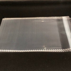May include: A stack of clear, transparent cellophane bags. The bags are rectangular with a stitched edge along the top. The bags are stacked on a black surface. The cellophane is thin and appears to be new.